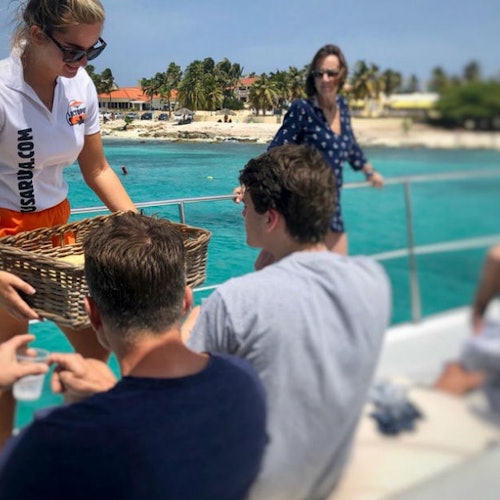 Un groupe de personnes sur un bateau ; une femme tend un panier à des individus assis avec une eau turquoise claire et une plage en arrière-plan.