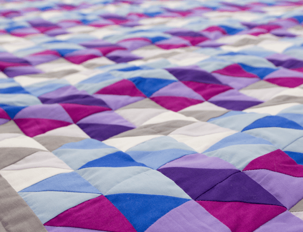 A close-up view of a quilt with a geometric pattern of interlocking triangles in shades of blue, purple, and pink. The quilt is composed of multiple fabric pieces stitched together, creating a repeating, wave-like design. The fabric pieces are arranged in overlapping rows, with some sections appearing lighter and others darker. The quilt is laid flat, filling the frame, and shows no visible edges or binding.