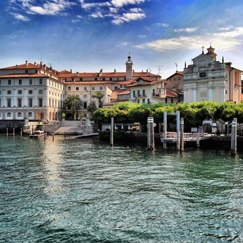 Waterfront view of historic buildings with red-tiled roofs, trees, and piers along a body of water under a blue, partly cloudy sky.