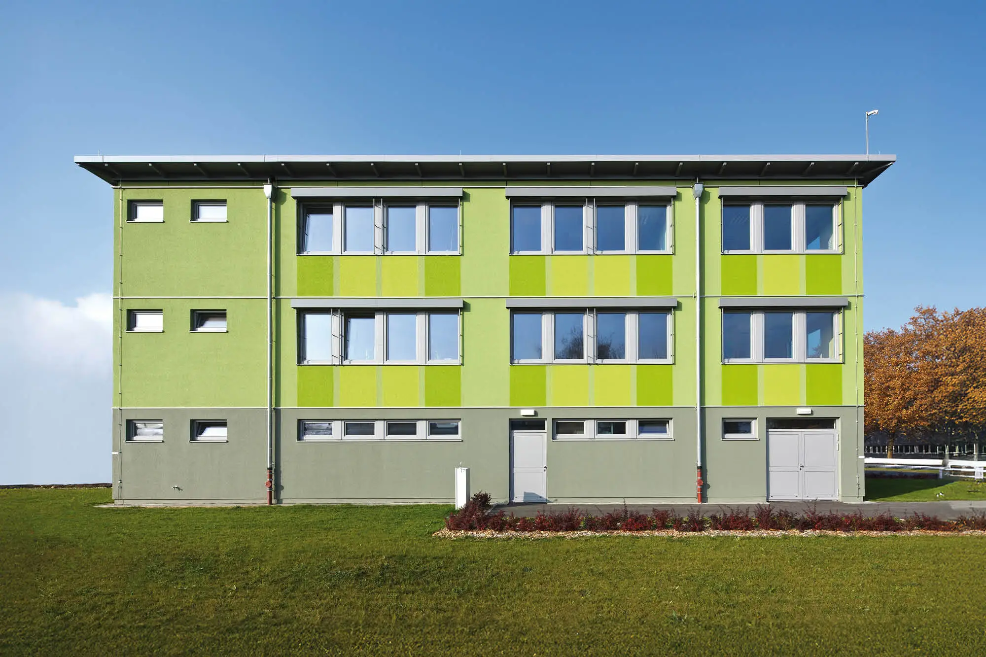 Grünes, dreigeschossiges Gebäude mit hellgrauem Sockel und vielen Fenstern, im Hintergrund Bäume mit Herbstlaub.