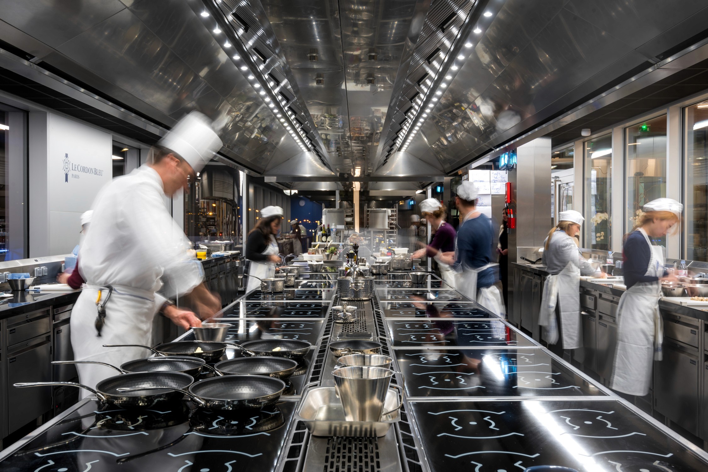 Chefs in white uniforms cook at a long row of stainless steel stoves and stations in a large industrial kitchen.