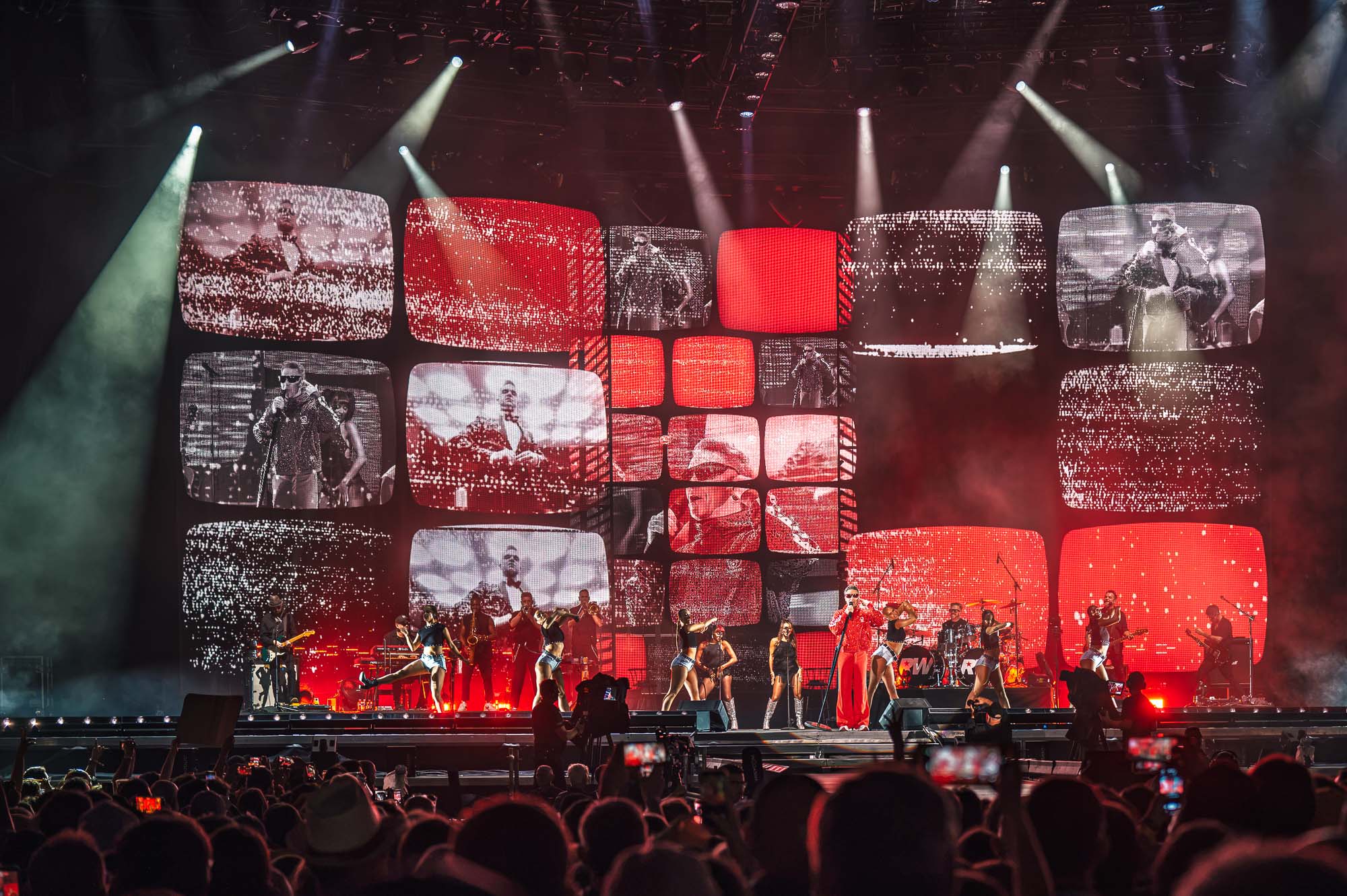 Mur vidéo façon grille de téléviseurs en rouge et noir avec images d’archives, danseuses et musiciens sur scène pendant le concert de Robbie Williams.