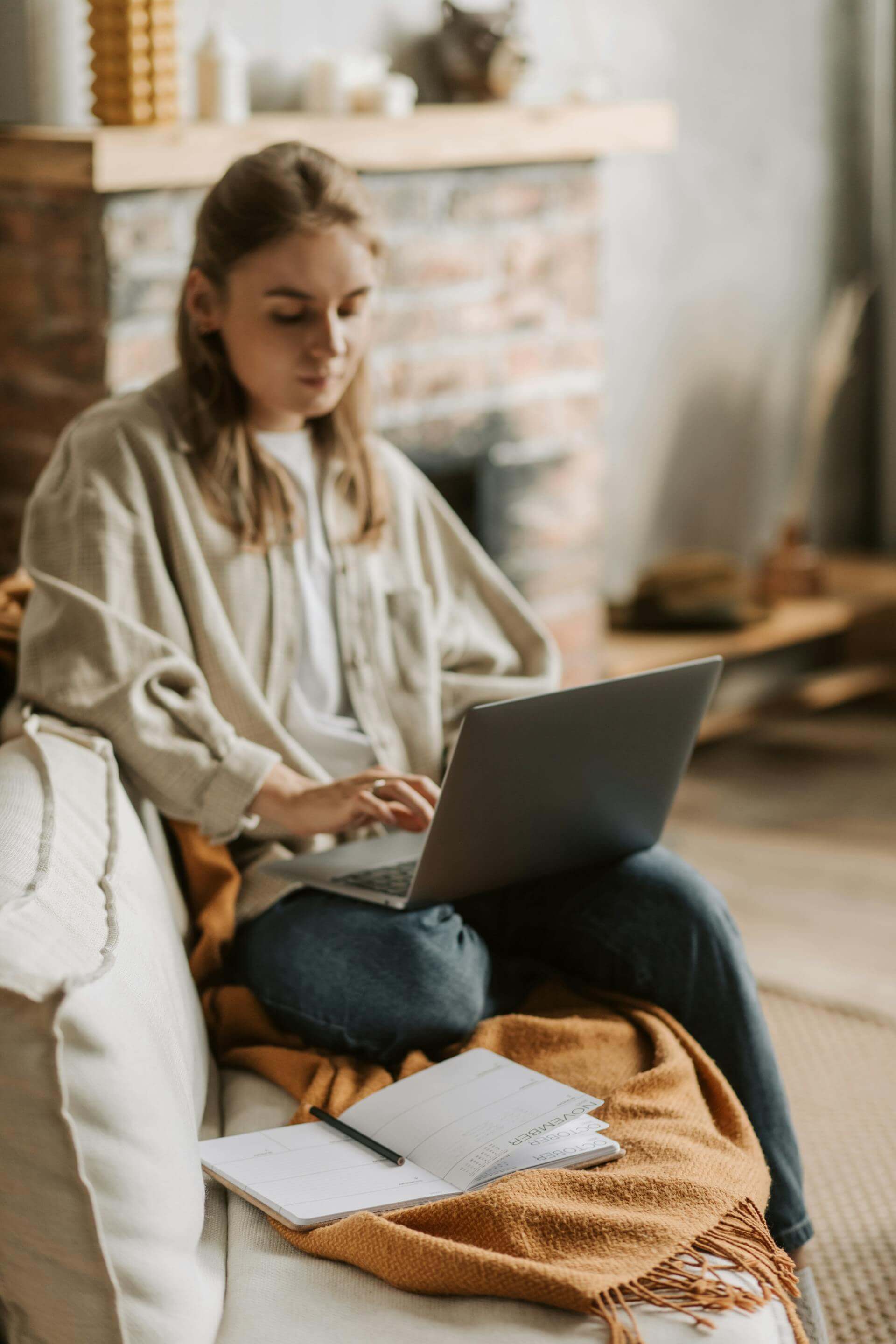 A woman seated on a couch, working on a laptop while taking notes in a notebook beside her