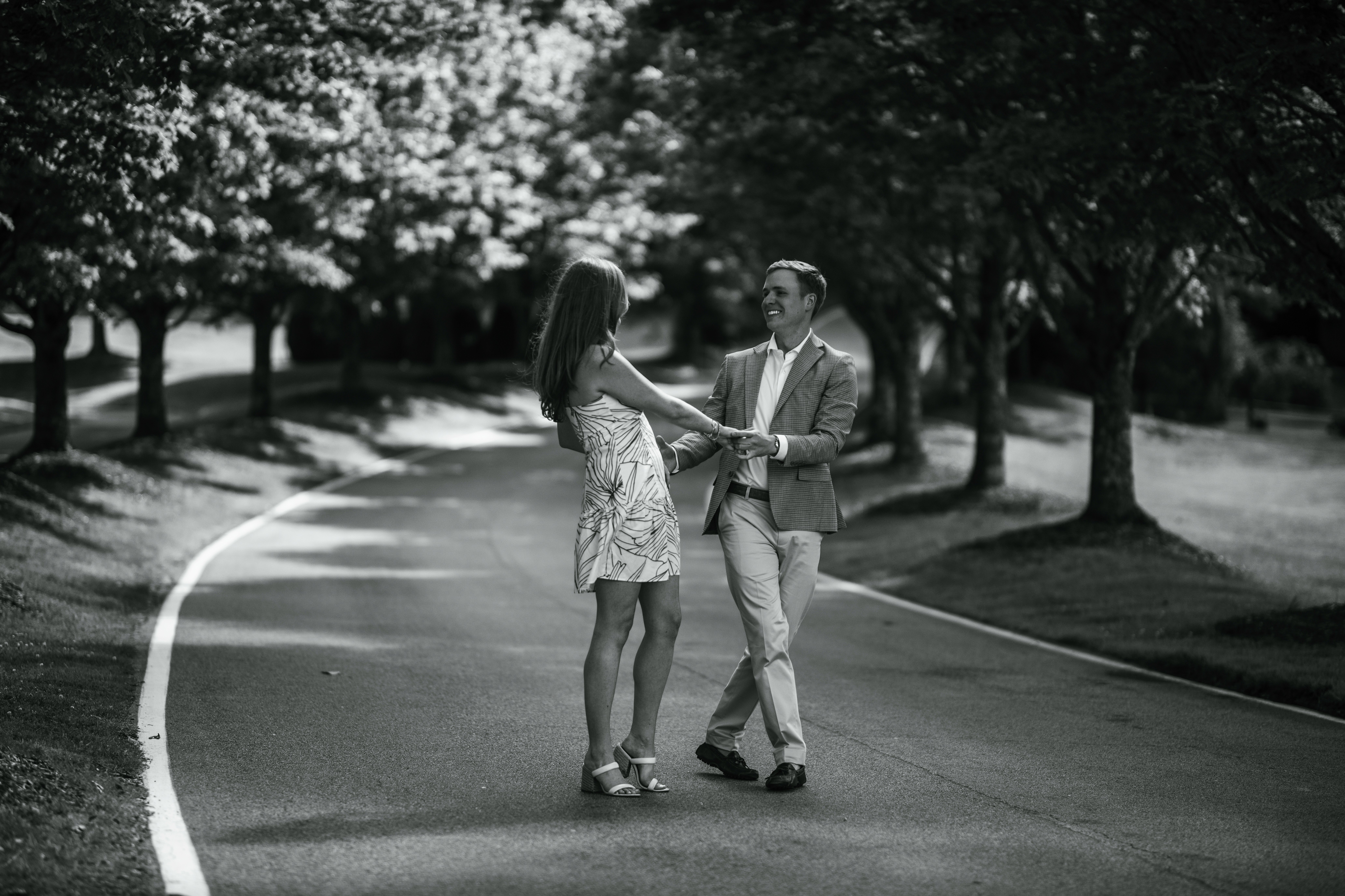 Couple dancing on a tree-lined road. Man in a blazer, woman in a dress and heels, smiling at each other.