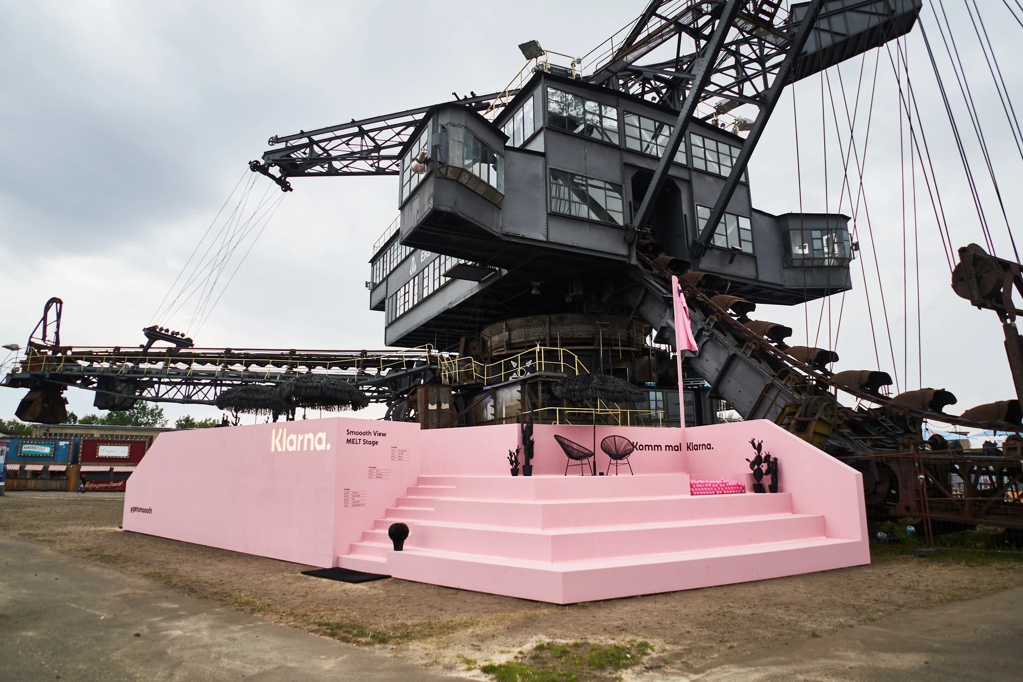 Pink Klarna „Smoooth View" stage at Melt Festival built in front of a massive industrial bucket-wheel excavator, with a flag and chairs on a raised platform.