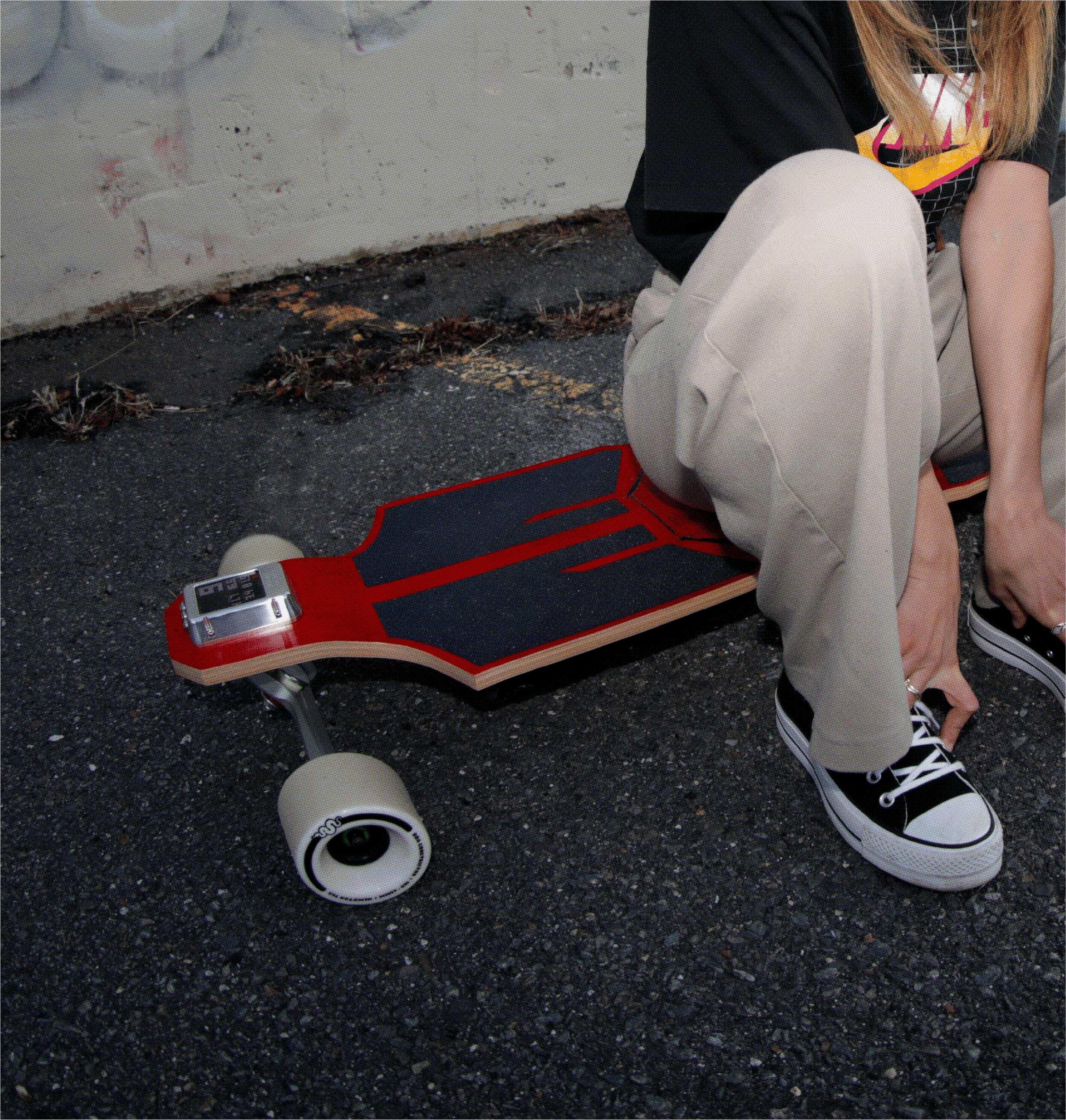 Person sitting next to electric skateboard on rough pavement wearing casual streetwear