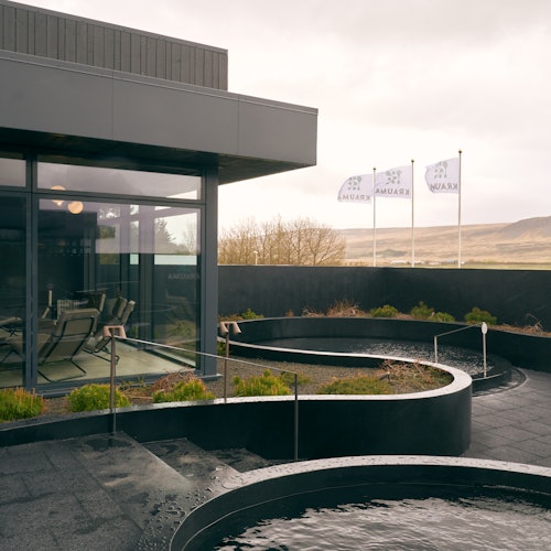 Outdoor modern patio with a glass-walled building, circular water features, plants, and several flags in the background.