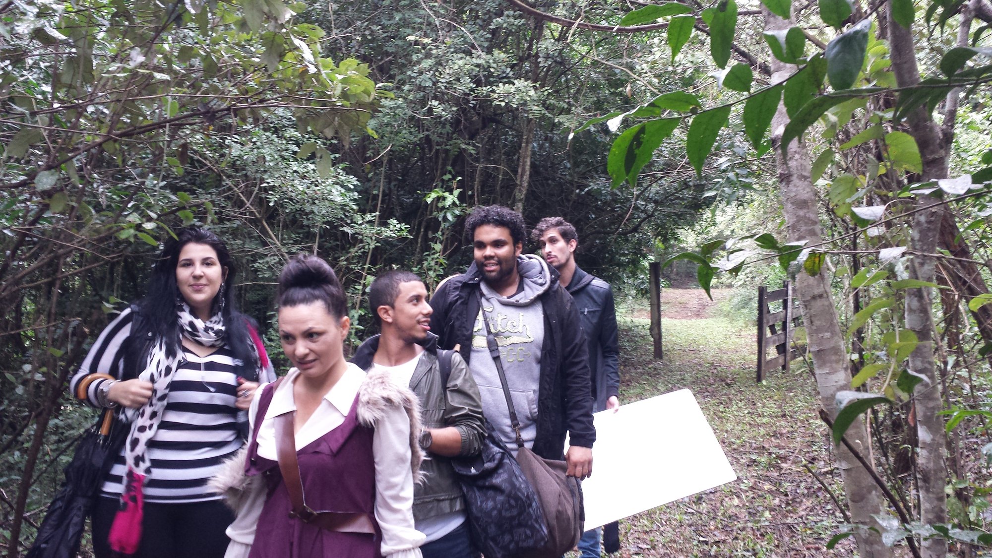 Grupo de pessoas caminha por trilha na mata carregando equipamentos, no making of com Vânia Leda