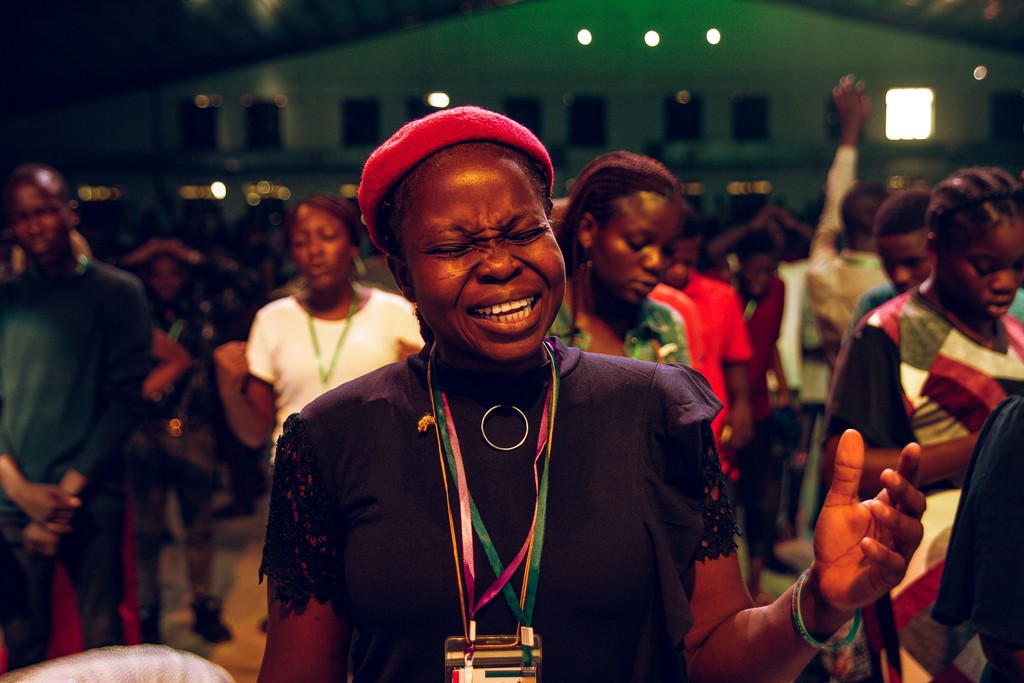 A woman passionately singing and worshiping during a lively, nighttime religious gathering, surrounded by other attendees engaged in deep prayer and worship, with vibrant lighting creating a spiritual atmosphere.