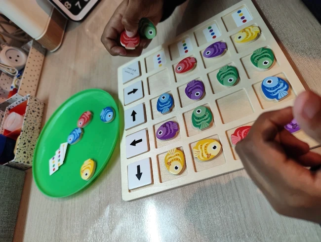 תקריב של ידי ילד פותר חידת התמצאות במרחב וקואורדינציה בעזרת לוח משחק מעץ עם דגים צבעוניים וחצים, כחלק מתהליך למידה חווייתית ופיתוח קוגניטיבי בקליניקה של סמדר עציץ - נעים ולומדים.