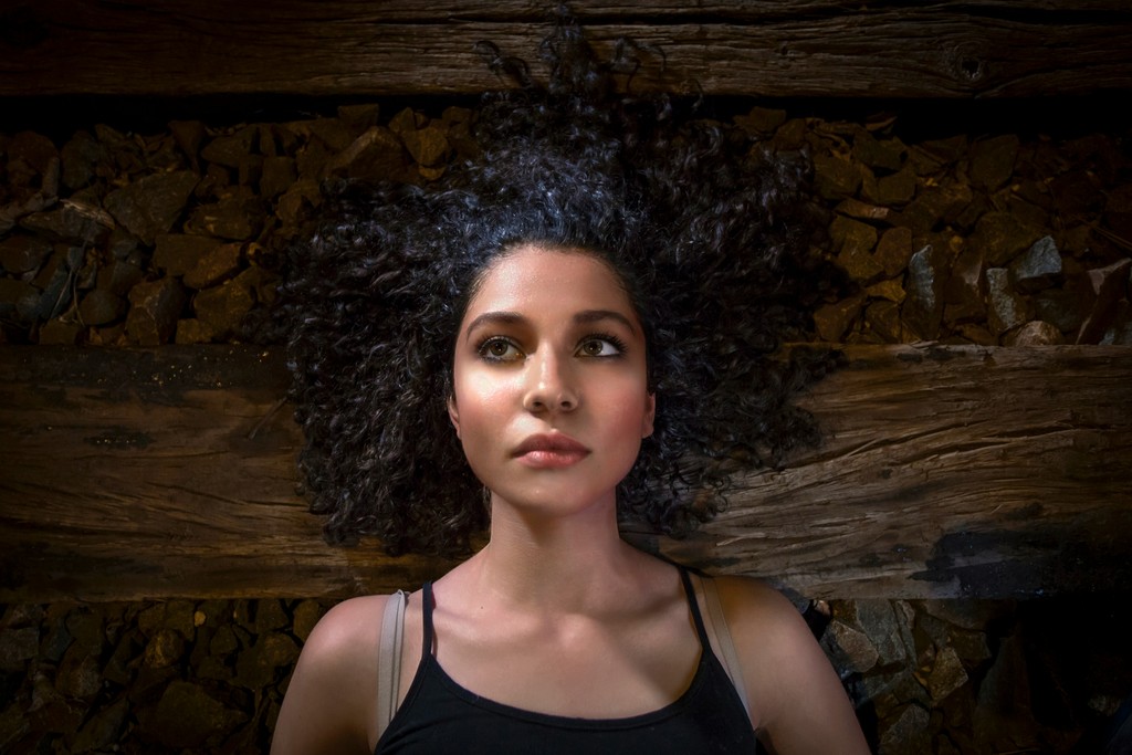 A woman with curly hair lies on a bed of rocks and wooden planks, gazing thoughtfully upward, evoking a sense of introspection and contemplation.