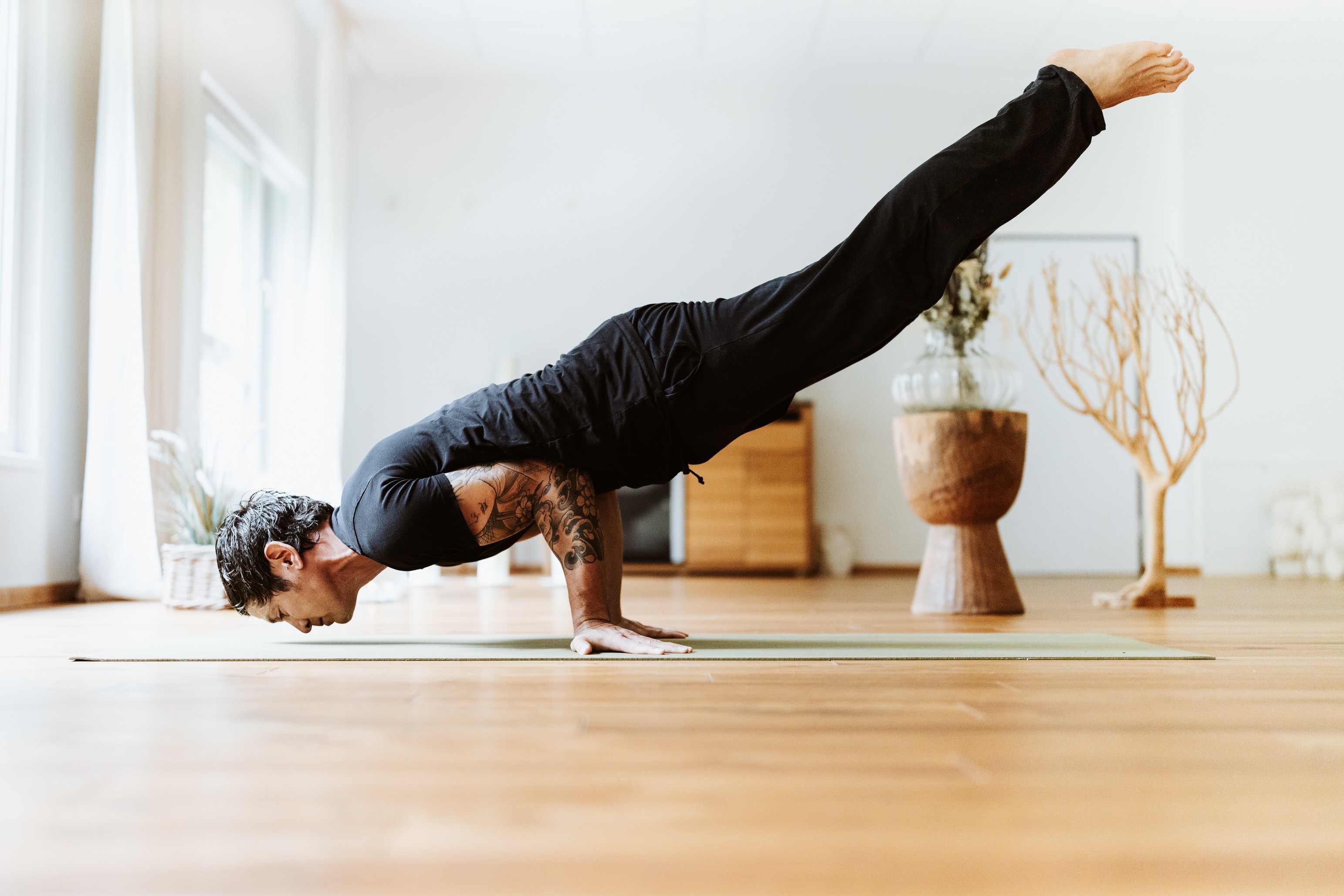 A person performs an advanced yoga pose, known as a Peacock Pose or Mayurasana, balancing on their hands with their legs extended horizontally in a serene, sunlit studio featuring wooden floors and minimalist decor.