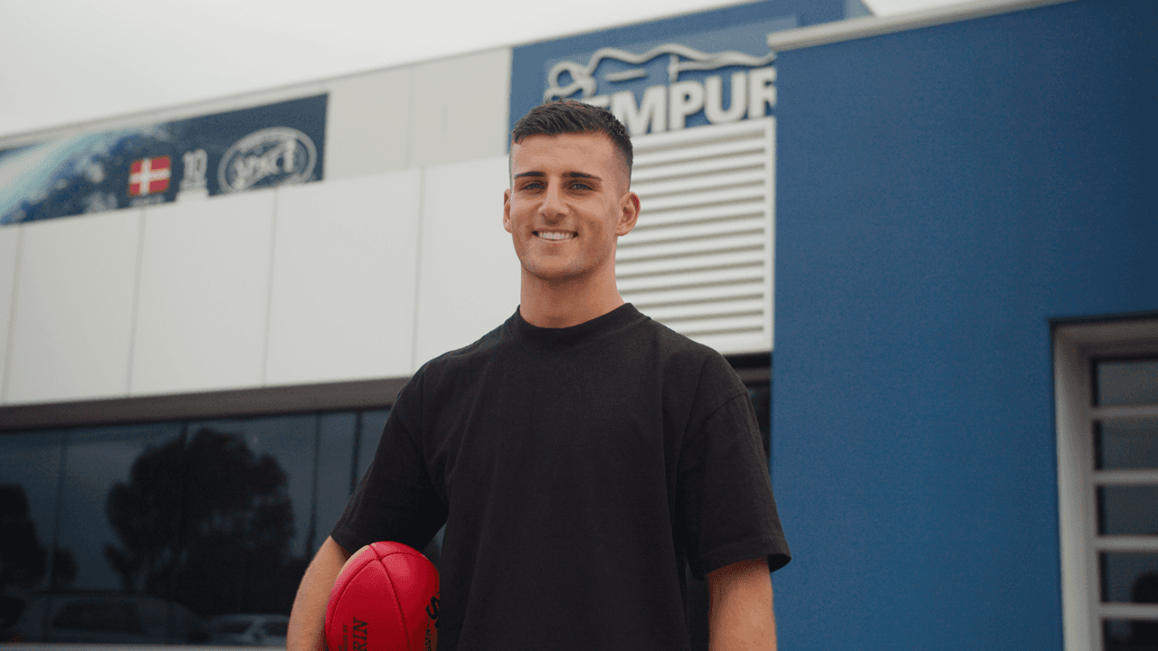 A young man with short dark hair wearing a black shirt stands in front of a blue background with what appears to be a boxing ring or training area visible behind him