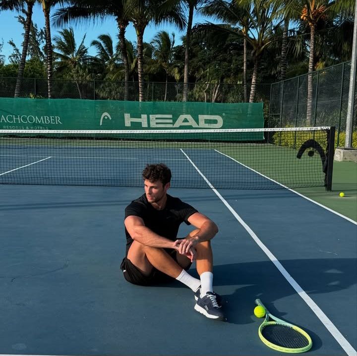 Smiling man adjusting the hood of his green and gray windbreaker, standing in a forest with tall bare trees in the background.
