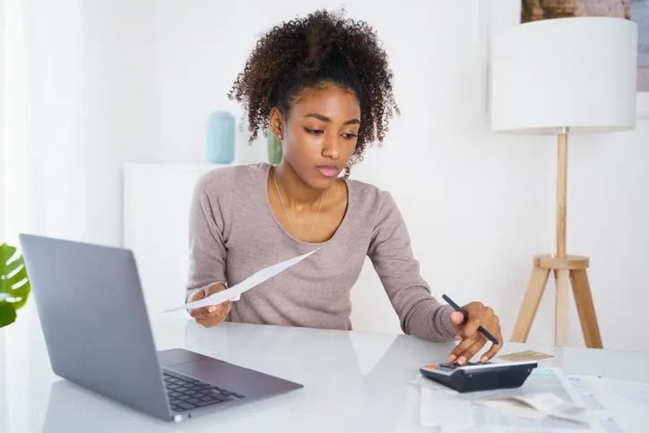 Young Black woman calculating finances on calculator with laptop and papers on desk