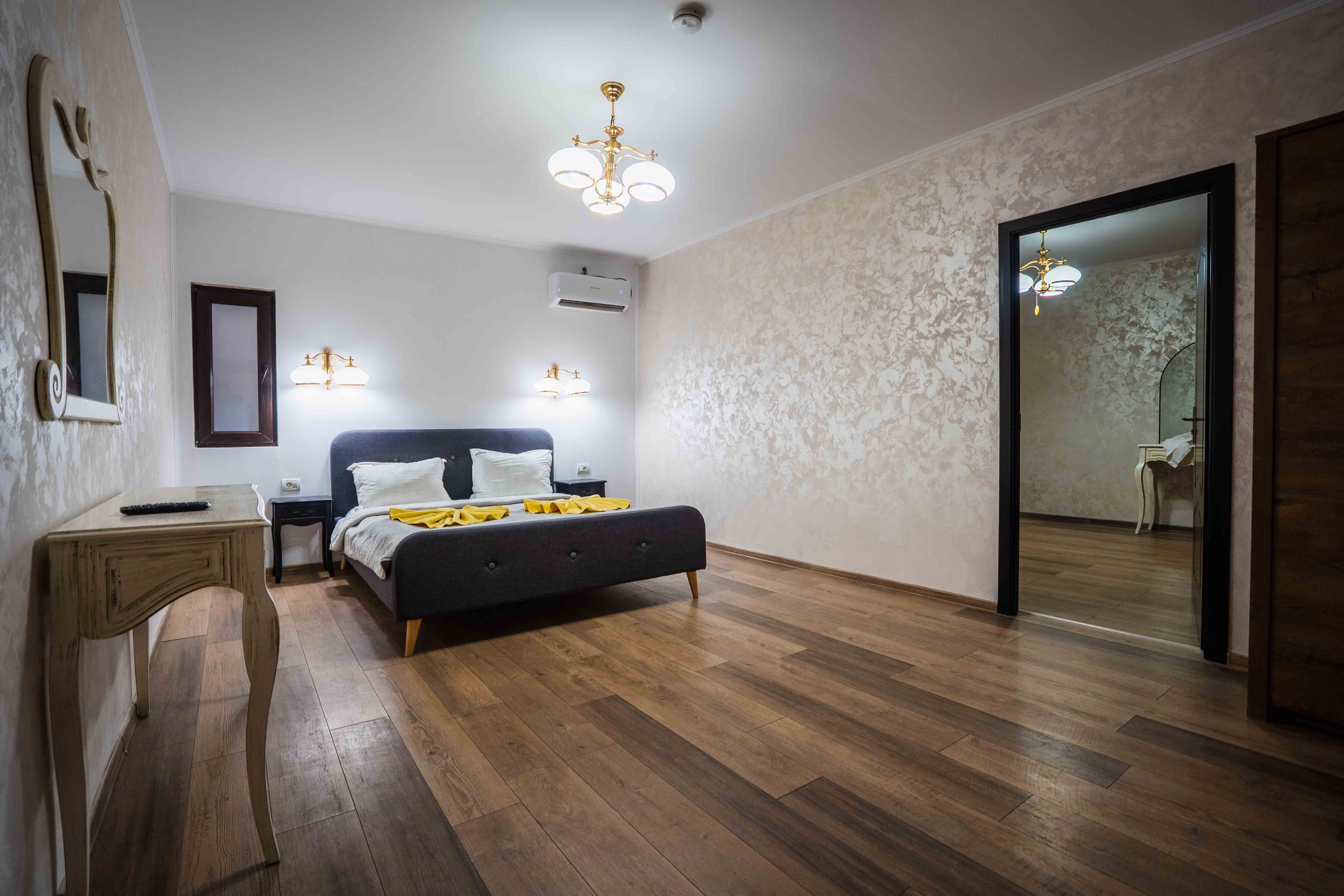 A modern, glass-walled house with warm interior lighting is perched on a rocky cliff overlooking a calm sea at sunset, under a clear blue sky.