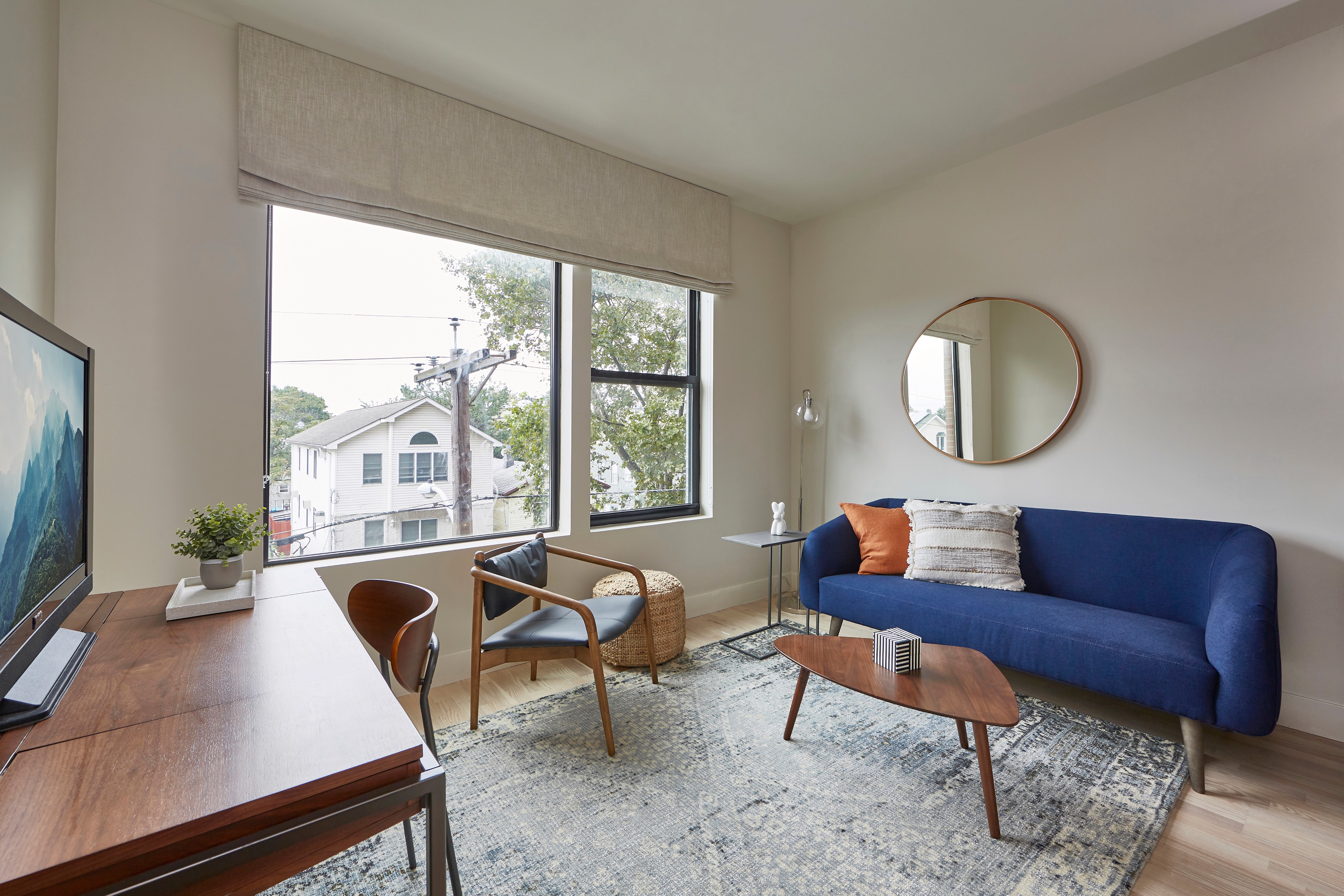 Cozy living room with a blue sofa, round mirror, and large window letting in natural light. Wood furniture accents.