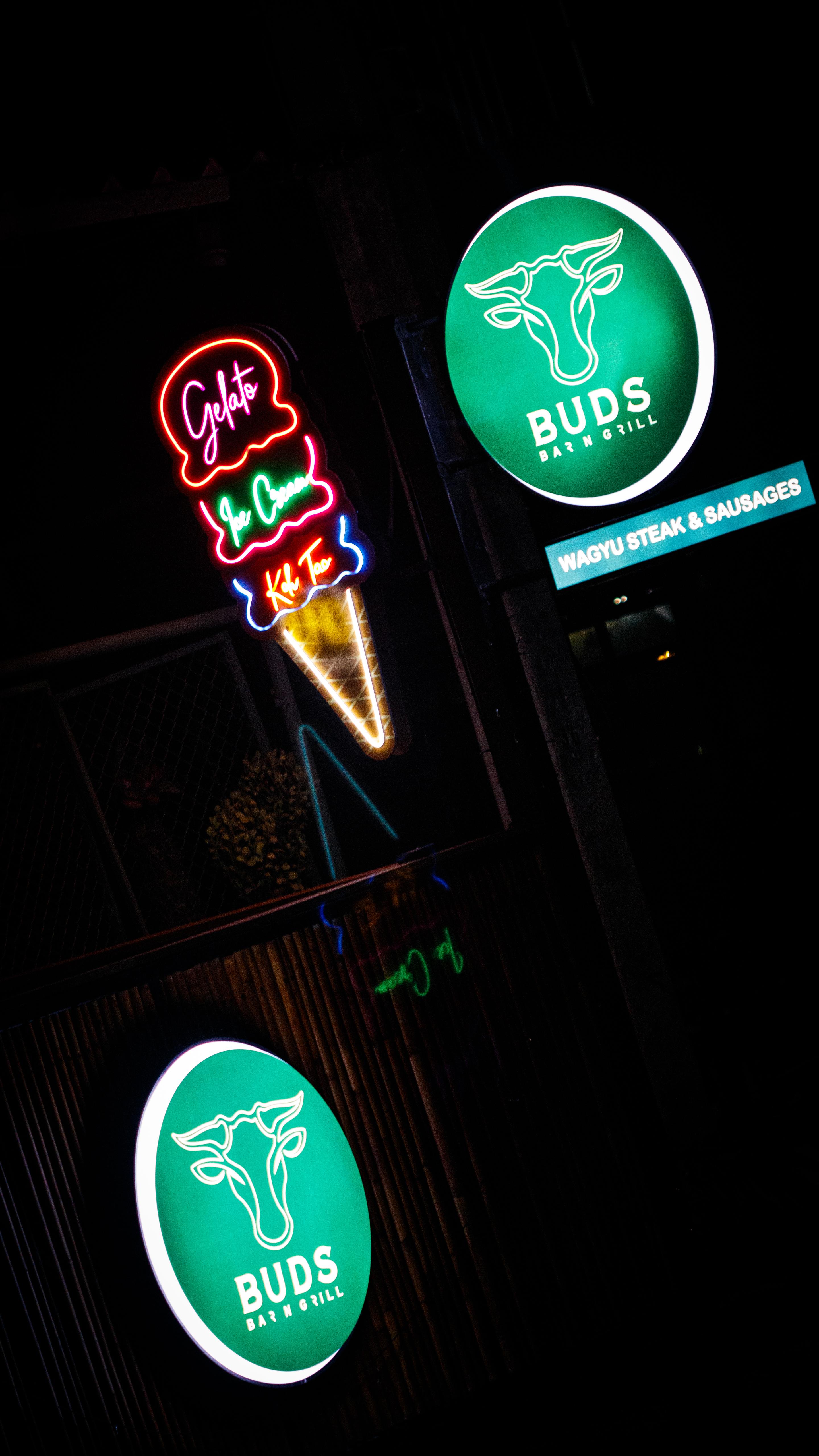A bright, modern dining room featuring a long light wood table set with several matching wooden chairs and white cushions, illuminated by natural light streaming through large windows overlooking a lush green and sunlit yard.
