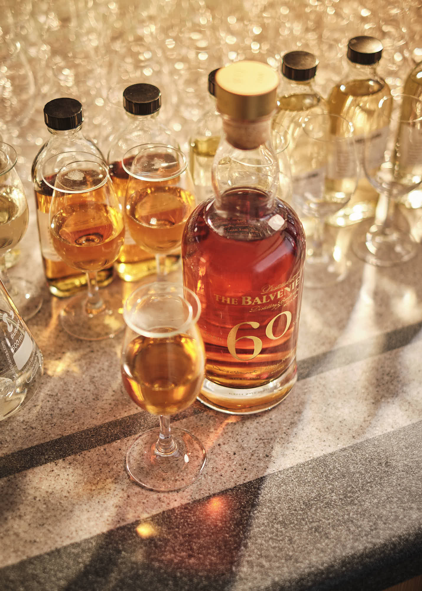 A close-up of a bottle of whiskey surrounded by glasses filled with amber liquid on a reflective surface.