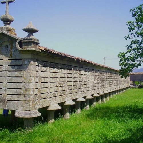 Un lungo e antico granaio di pietra su pilastri rialzati in un campo erboso, sotto un cielo azzurro e limpido con un piccolo albero sul lato destro.