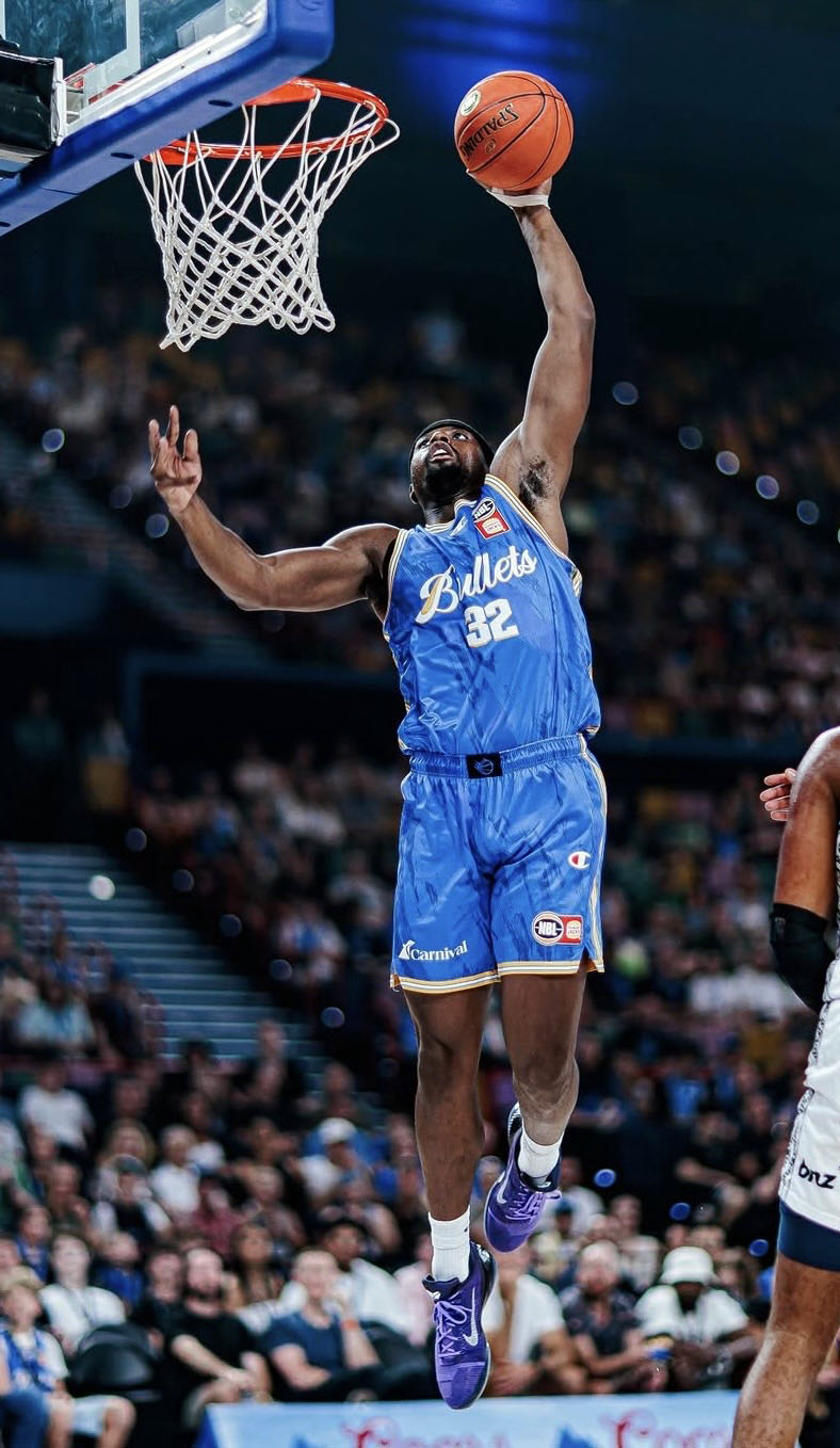Terry Taylor in a blue Bullets uniform leaps towards the hoop for a dunk during a game, with a crowd watching in the background.