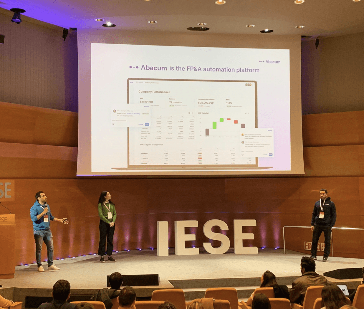 A presentation at IESE with three speakers on stage, discussing a topic with a visible slide in the background.