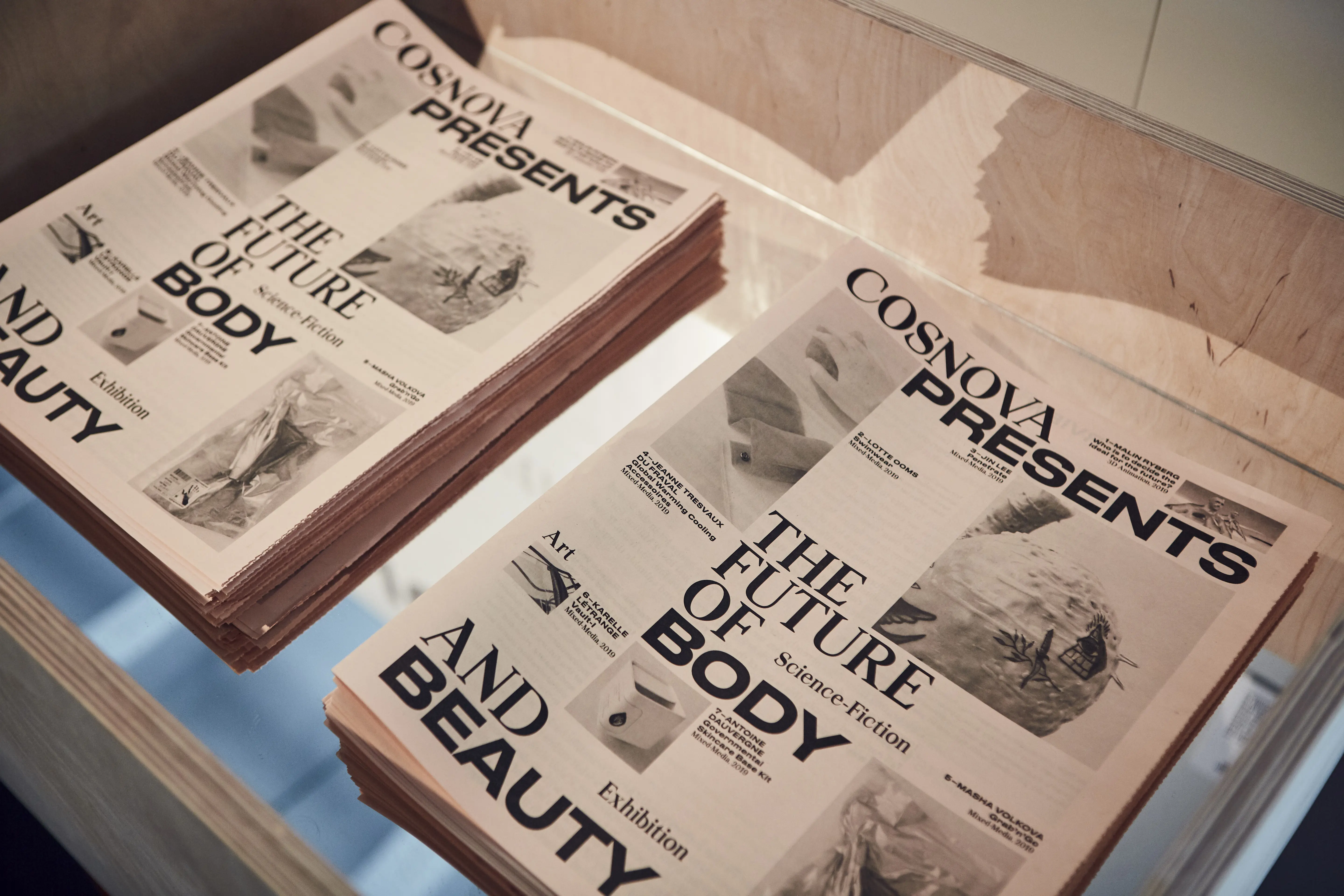 Stacks of the exhibition newspaper Cosnova Presents The Future of Body and Beauty on a plywood display table.