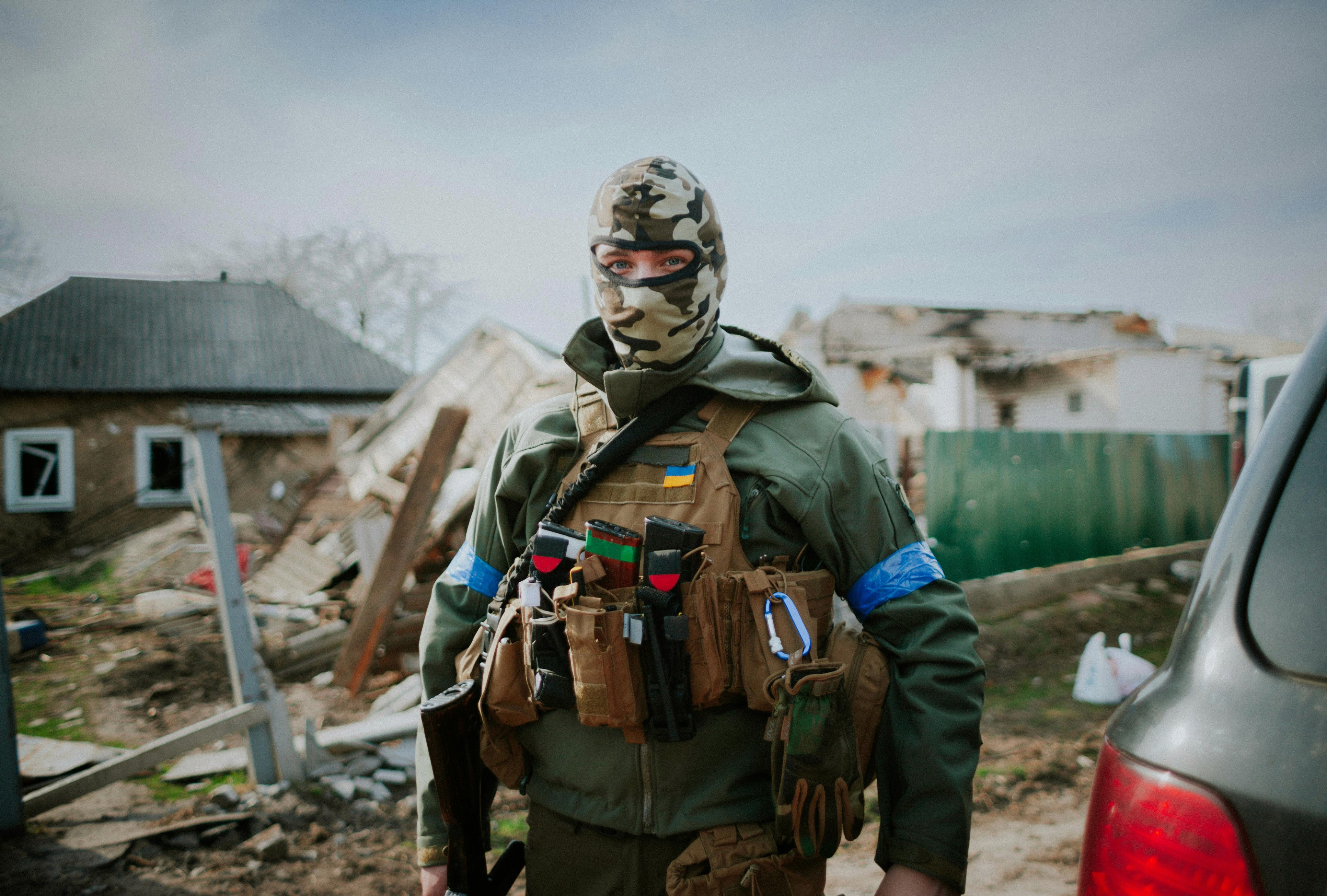 Ukrainian soldier with only his eyes visible, wearing a military jacket, cap and scarf, with a backpack and a Ukrainian flag patch on his shoulder; standing in front of destroyed buildings.