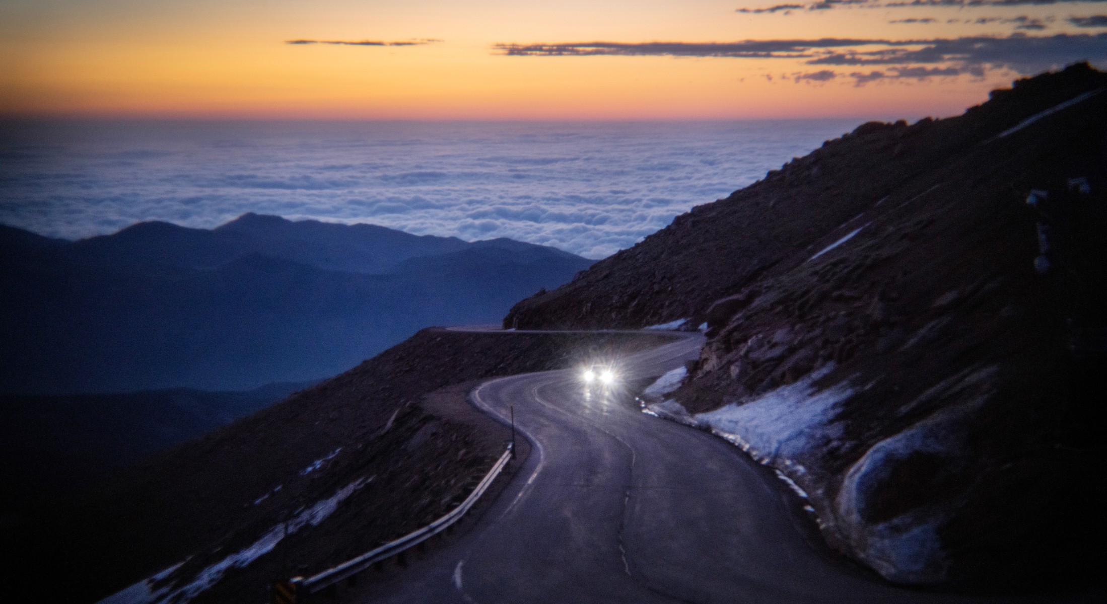 A race car with its headlights on, driving up Pikes Peak at dusk.
