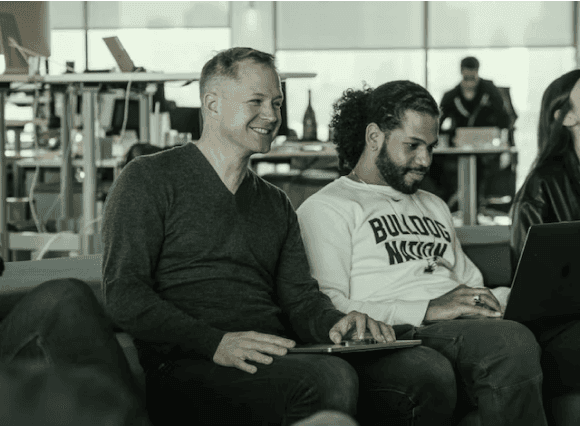 Two people are comfortably seated on a couch, each with a laptop, collaborating on tasks in a relaxed environment.