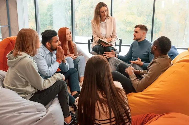 A circle of people sitting cross-legged, meditating with hands in a prayer position. Soft, warm lighting.