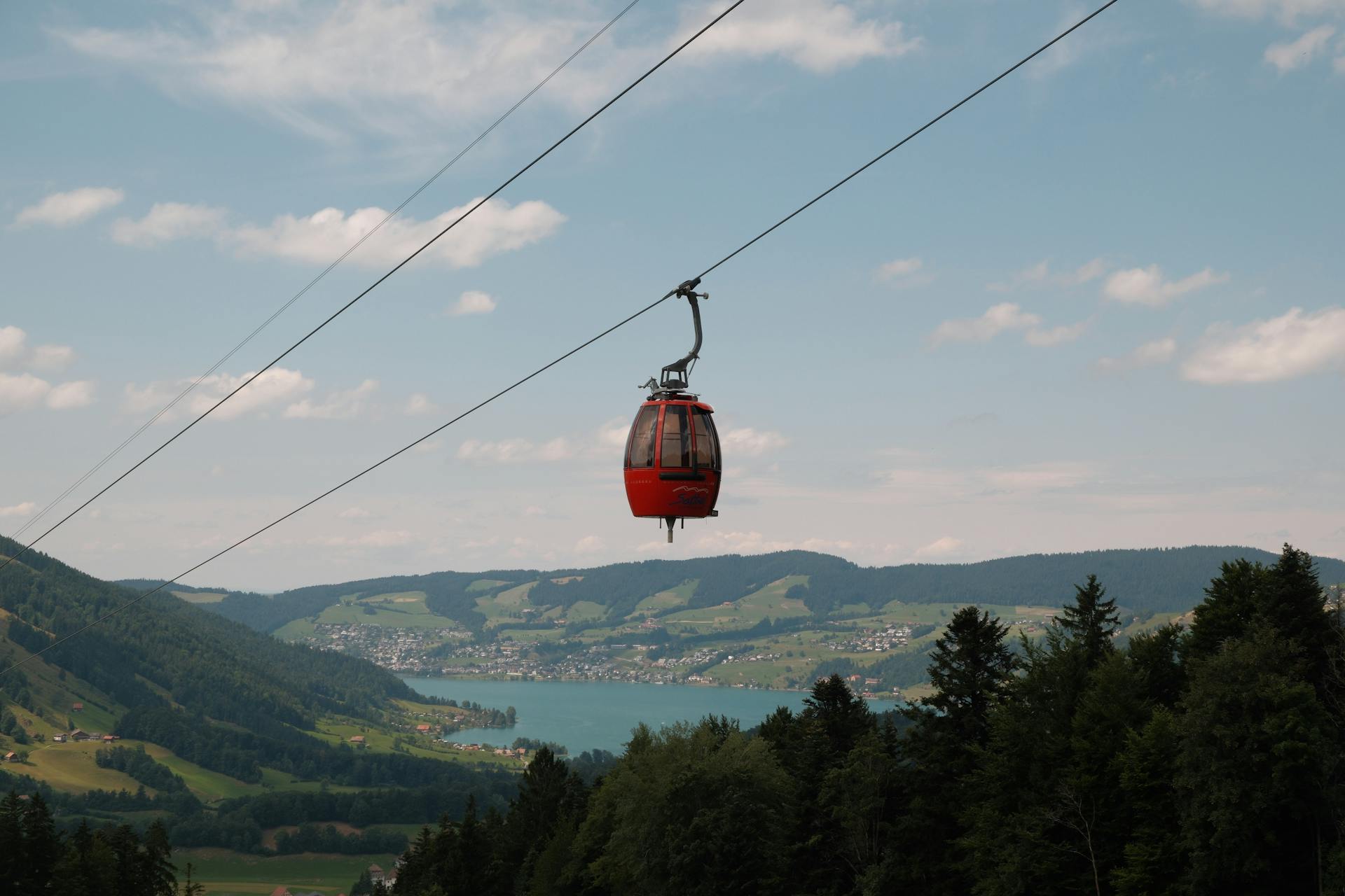 A red gondola car travels on cables over a green valley with a large lake and a town on its shore.