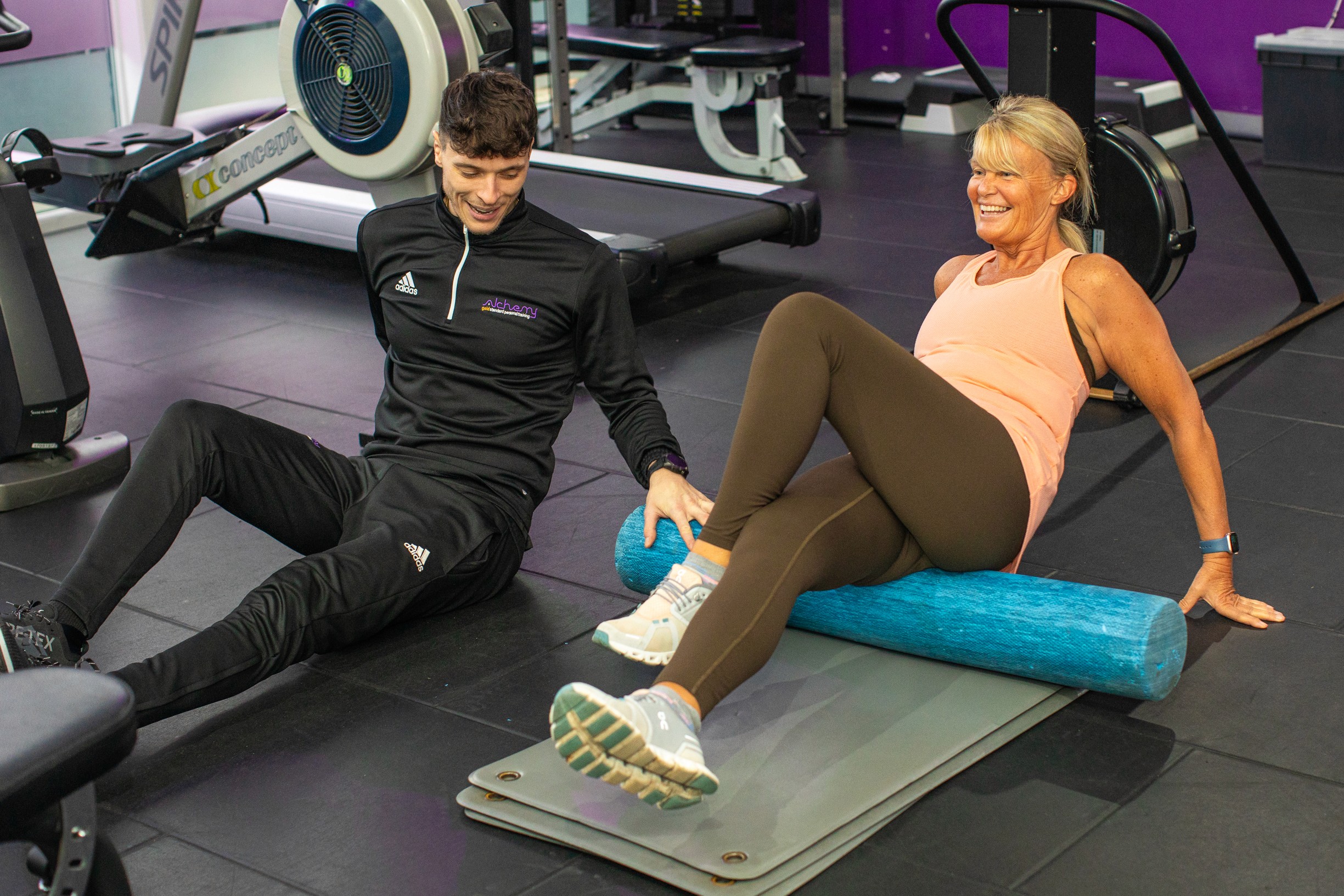 A personal trainer is coaching a woman through a foam rolling exercise on a mat in a gym, with cardio equipment in the background.