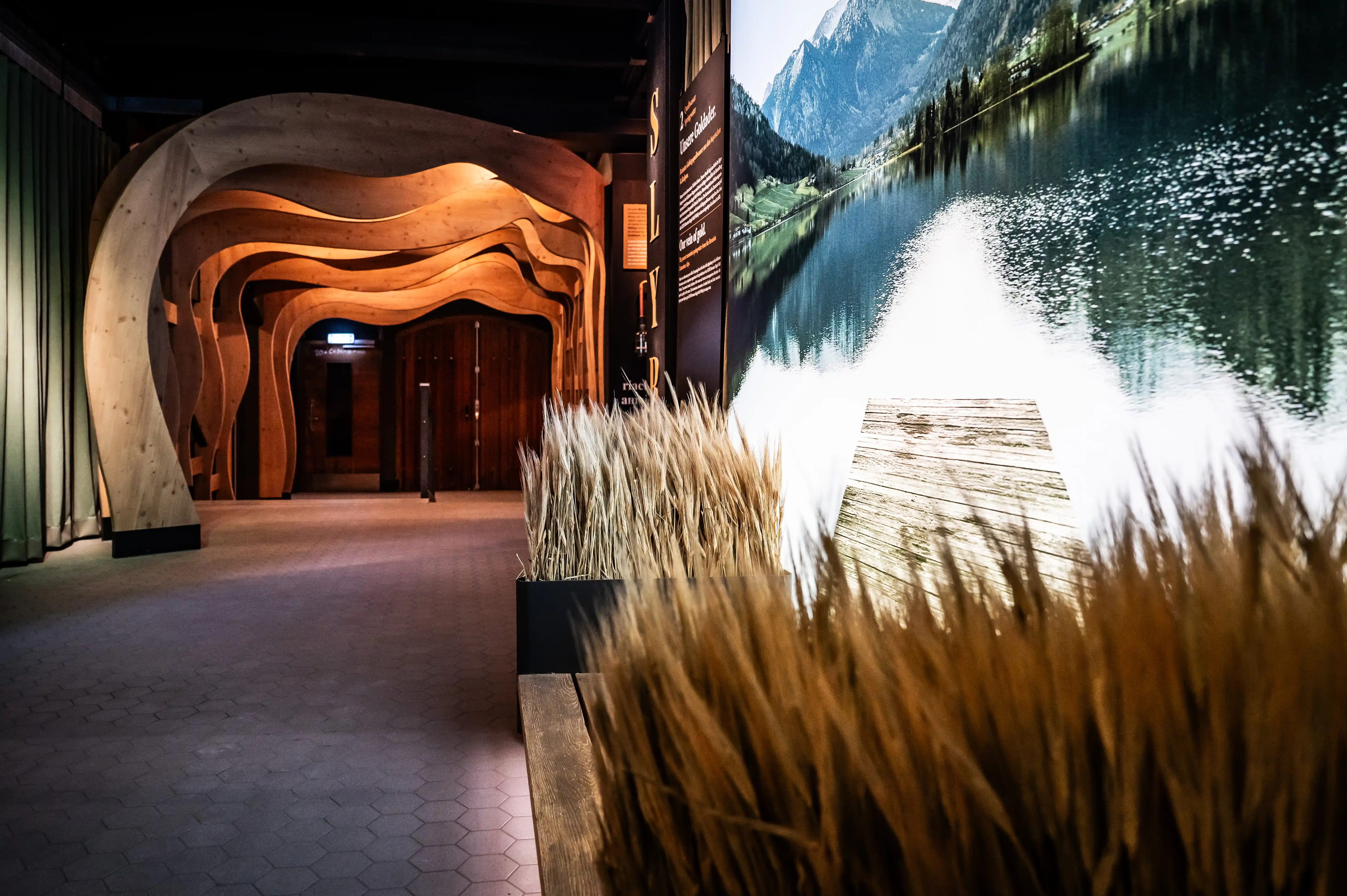 Organically curved wooden tunnel leading through the SLYRS Experience Centre past a glowing mountain-lake panorama.