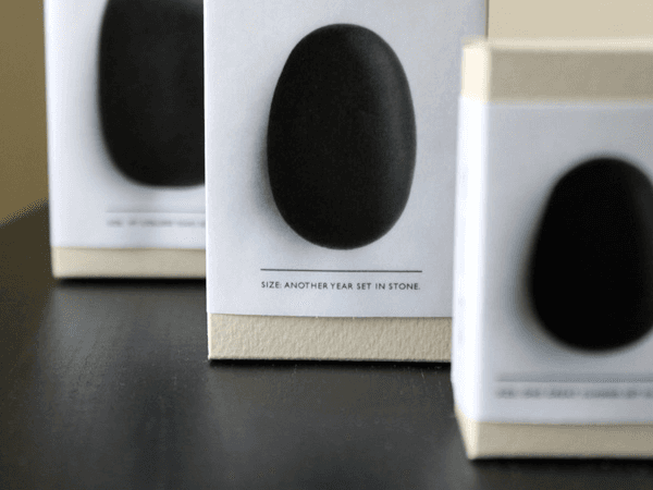 Three black oval stones are displayed on white boxes. Each box has a black oval image and the text "SIZE: ANOTHER YEAR SET IN STONE." printed below. The boxes are arranged on a dark surface.