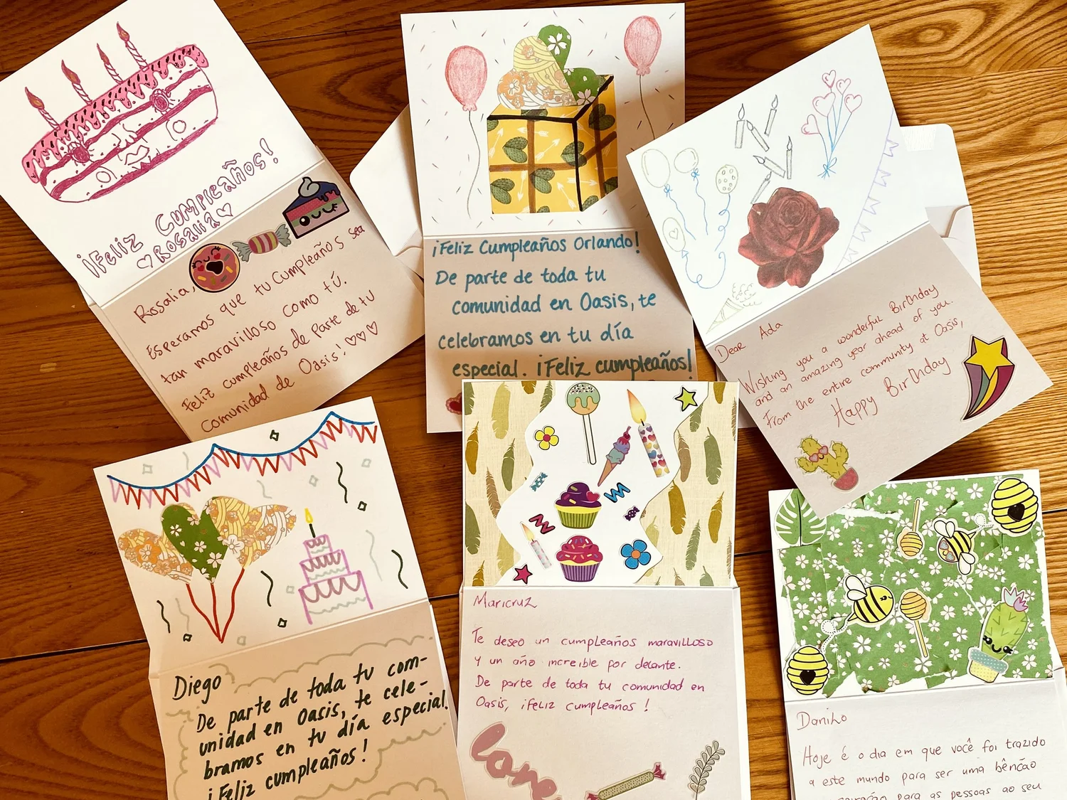 Uma coleção de cartões de aniversário feitos à mão com decorações coloridas, apresentando desenhos de bolos, balões e presentes de aniversário, dispostos sobre um piso de madeira.