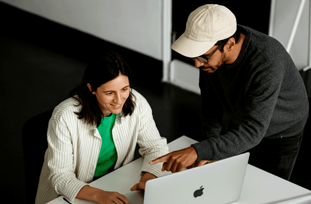 Two people looking at a laptop, one points at the screen. Man wears a cap and glasses. Woman in a green shirt.