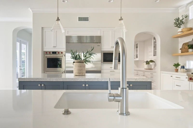 View over quartz sink to blue island, pendant lights, and subway tile in Bonita Canyon Remodel.