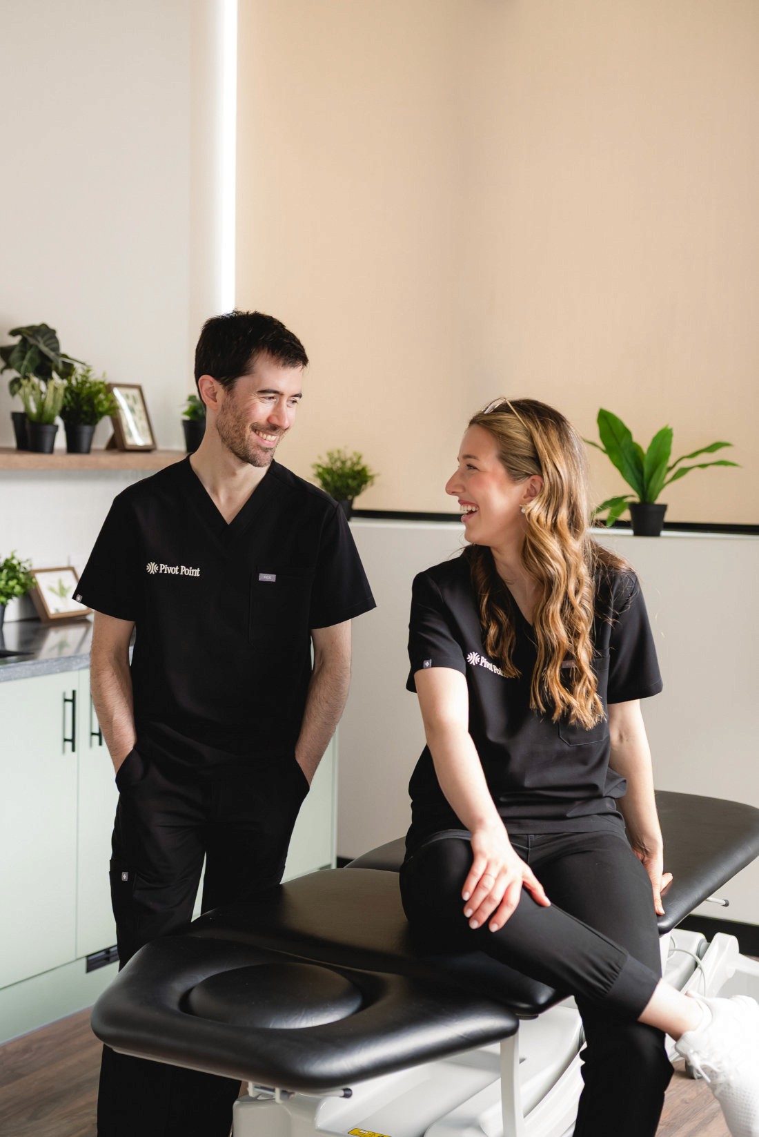 Two physiotherapists in uniform in a treatment room, facing each other and smiling