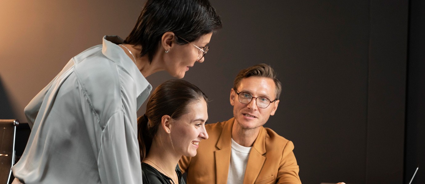 Three people collaborate around a table in a dimly lit office, with one person pointing at a laptop screen, indicating teamwork and discussion.