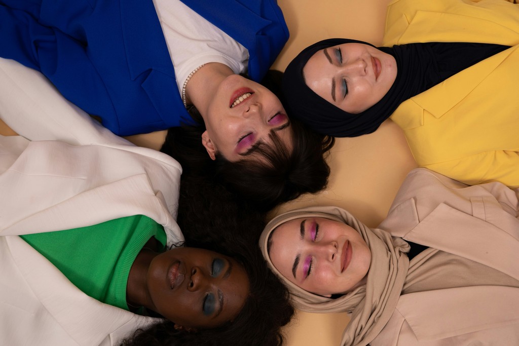 A diverse group of four women, dressed in vibrant outfits, lie on the floor with their heads together in a circle against a beige background. They all have their eyes closed, showcasing colorful eye makeup, and their expressions are peaceful and content, reflecting unity and harmony.