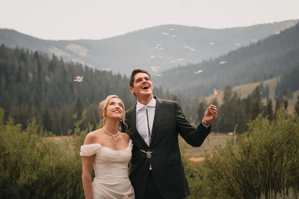 A playful moment during a Montana ranch wedding where the bride and groom look up and laugh as dandelion seeds float in the air around them against a mountain backdrop.