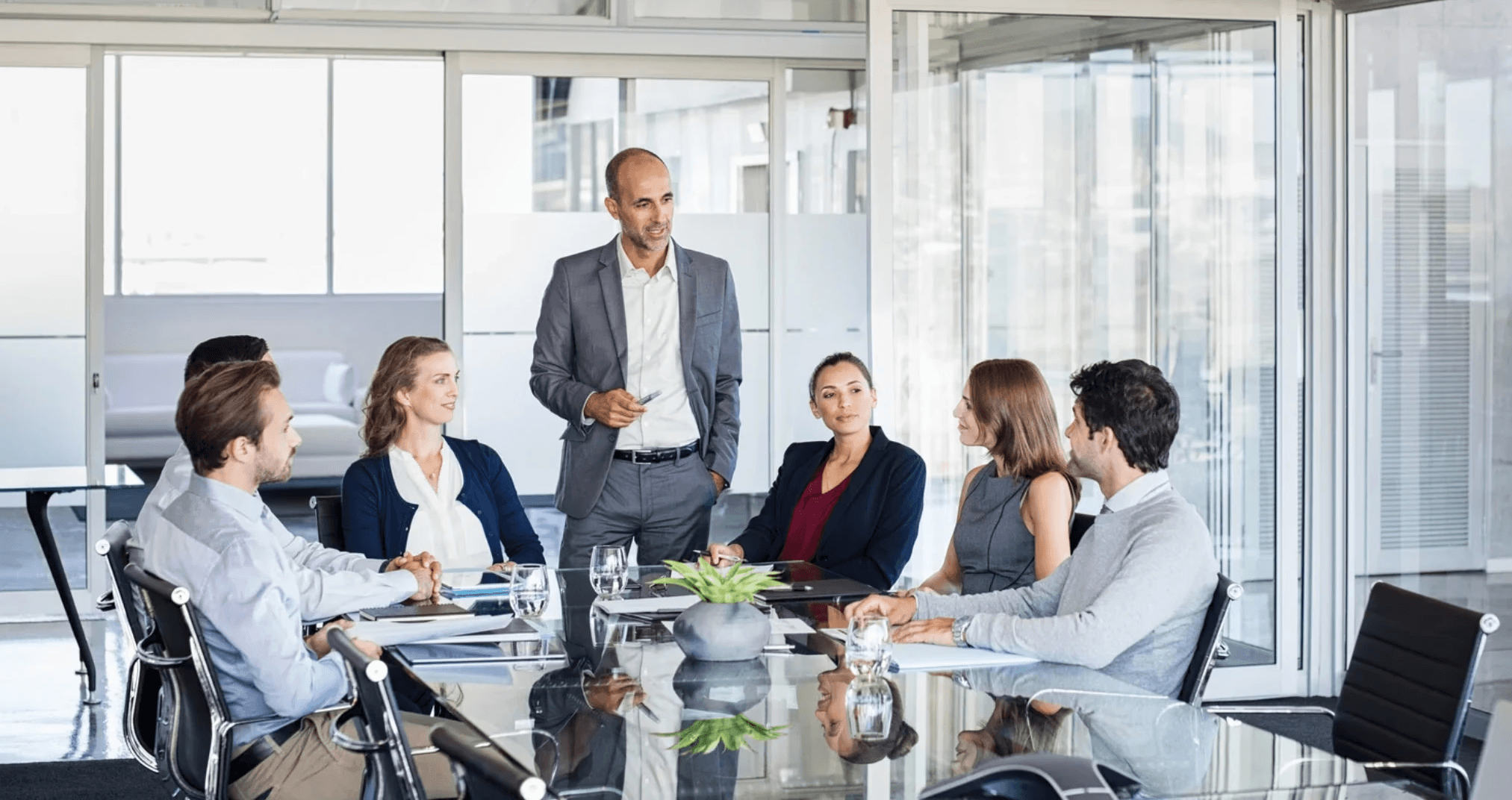 Geschäftsleute in einem modernen Konferenzraum während einer Besprechung, ein Mann steht und präsentiert vor einem Team am Tisch.