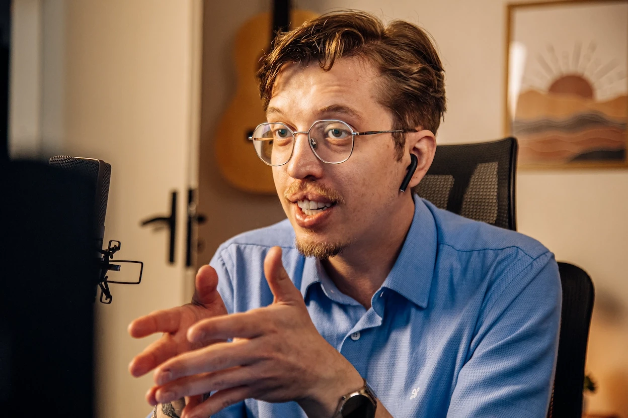 Homem de óculos e camisa azul, com fone de ouvido, gesticula em frente a um microfone, possivelmente gravando um podcast ou videochamada, com violão e quadro de paisagem no fundo.