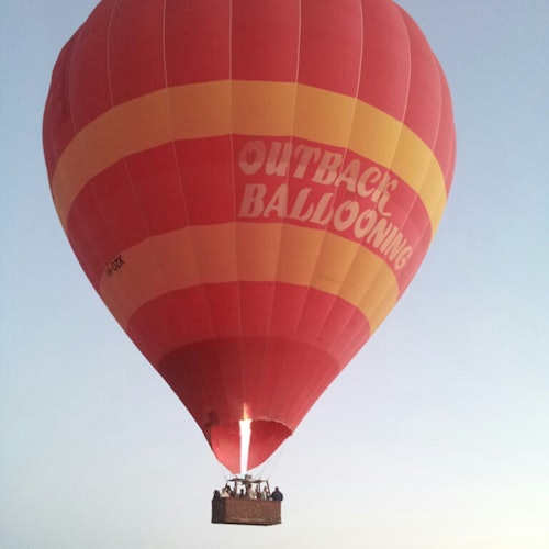 Red and yellow hot air balloon with "Outback Ballooning" text, carrying people, rising against a clear sky.