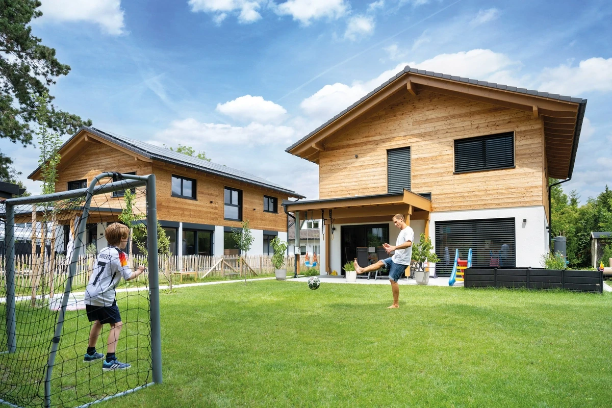Ein Mann und ein Junge spielen Fußball im Garten vor zwei modernen Holzhäusern bei strahlend blauem Himmel.