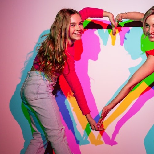 两个人用手臂在白墙上形成心形，投射出五彩斑斓的阴影。
