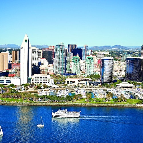 Horizonte de la ciudad con varios edificios altos, un parque junto al río y barcos en un río azul bajo un cielo despejado.