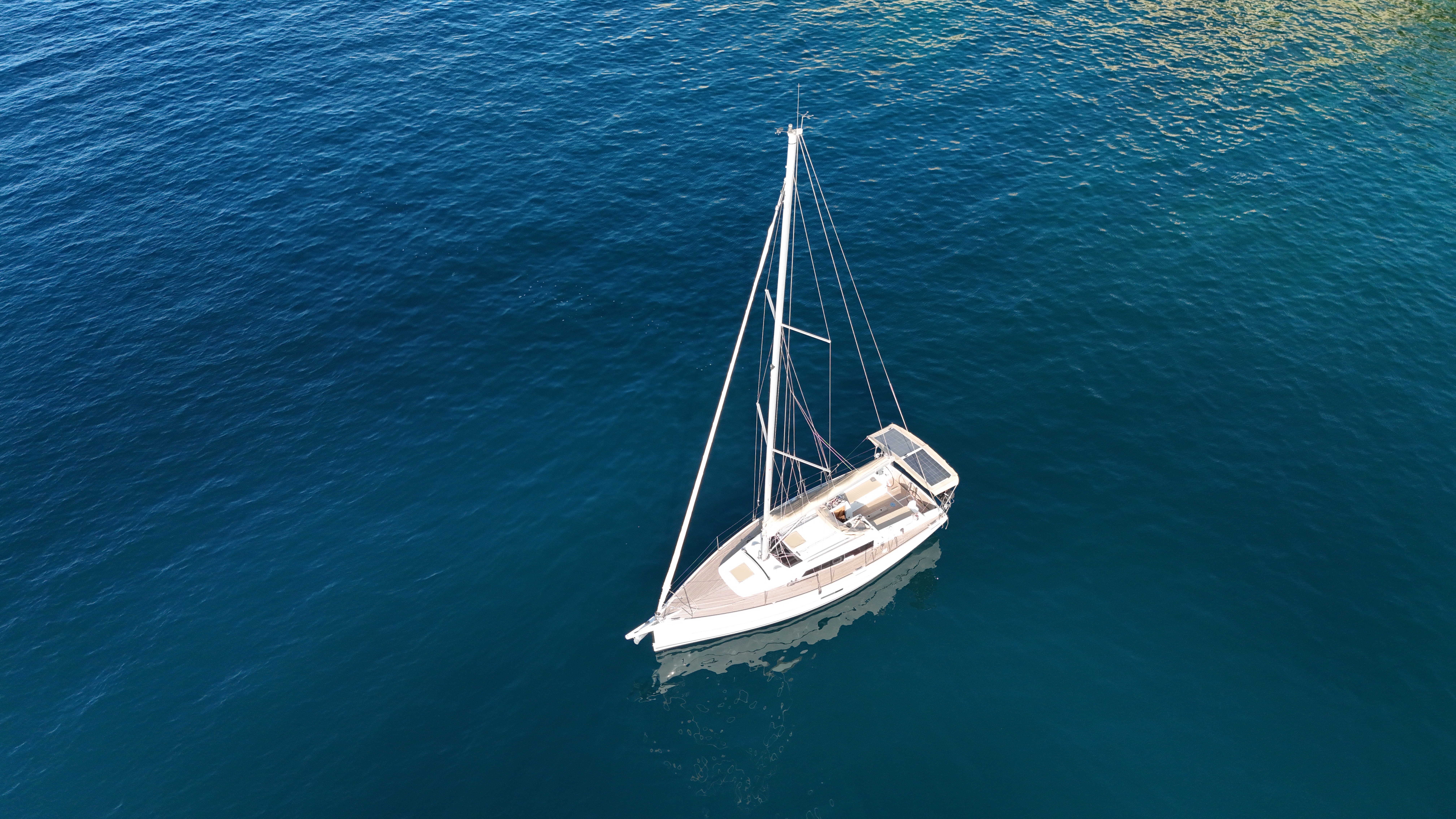 Aerial view looking down on a Dufour 350 GL sailing yacht anchored in deep blue water, showing its teak-accented deck, solar panel over the cockpit, and tall single mast.