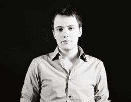 A black and white portrait of a man against a black background. He is wearing a light-colored, long-sleeved button-down shirt. His hair is short and dark, and he has a neutral expression. The lighting is focused on his face.